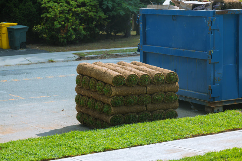 Sod is stacked next to a blue dumpster, and is ready to be laid out in the yard.